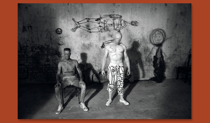 Black and white photograph of two men, one sitting, one standing in a cemented area surrounded by some wire sculptures