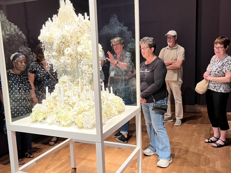 A variety of white people look at the Zelda sculpture in its glass case. The sculpture is covered in white wax, has a vaguely human form, candles, beads, and tiny wax hands cover it.