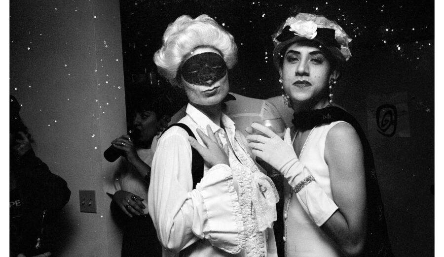 Black and white photo of two androgynous people with white skin, wearing hats, one wearing a mask, appear to be at a costume party.