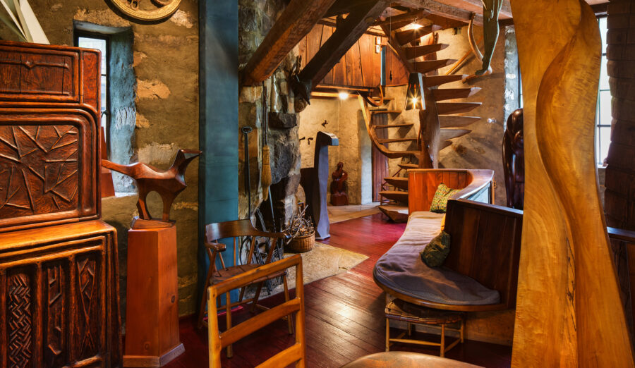 A cluttered room featuring a handmade spiral staircase and many other handmade carved wood furnishings