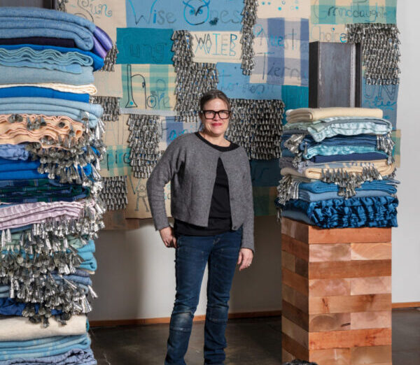 The artist in her workspace surrounded by piles of blue fabric and a black dog.