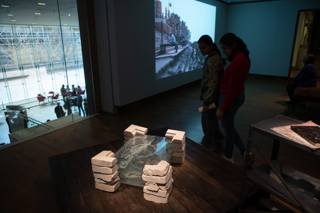 A darkened room with a window looking out into the lobby, a film projected on the wall, two people looking at a stacked series of engraved rectangles of glass supported by remnants of square concrete forms.