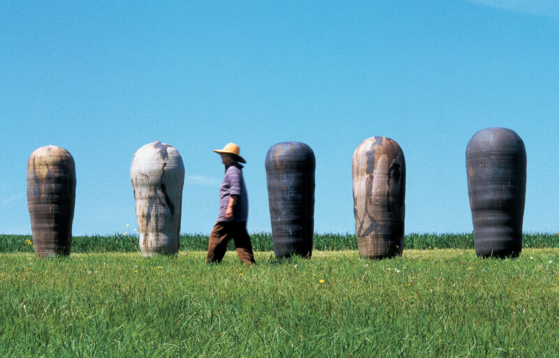 A green field, blue sky, with enormous ceramic vessels on the horizon. The artist, who is about the same height as the works, walks in front of them
