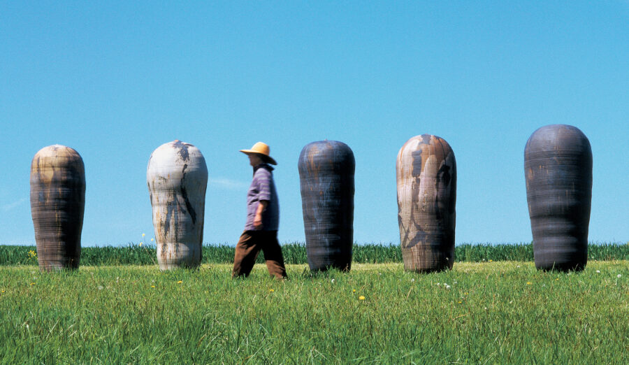 A green field, blue sky, with enormous ceramic vessels on the horizon. The artist, who is about the same height as the works, walks in front of them
