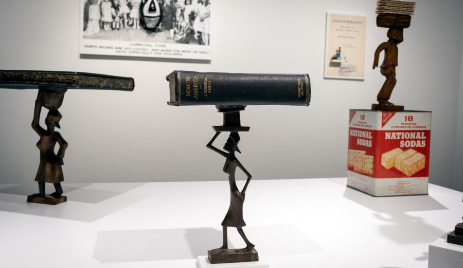 a stylized statuette of a woman apparently carved in dark wood is topped by a large heavy book