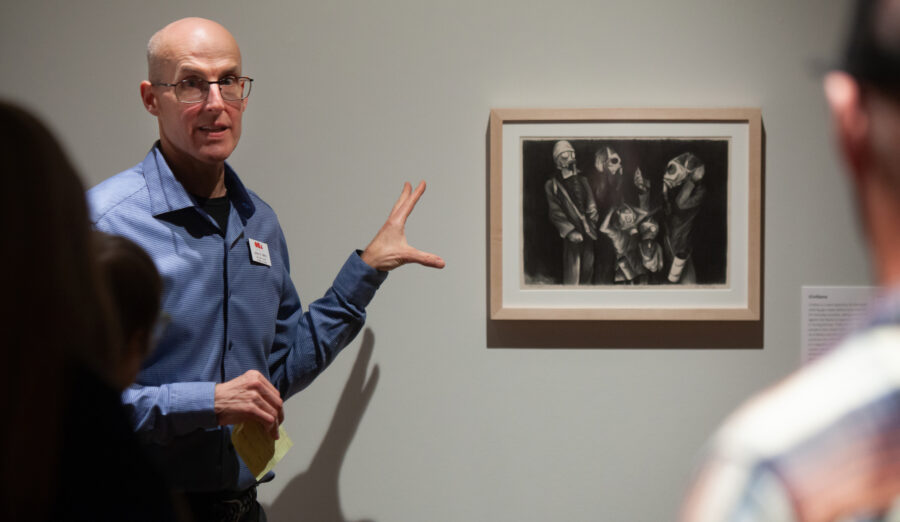 A person gestures toward a framed black-and-white artwork on a gallery wall while speaking to an audience.
