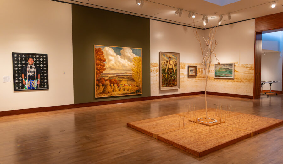 A spacious art gallery with wooden floors and multiple framed paintings on the walls, centered around a minimalist wooden platform with a bare tree and small circular objects.
