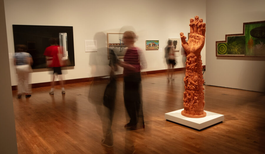 A blurred motion photograph captures visitors walking through an art gallery, with a prominent orange hand sculpture on display.