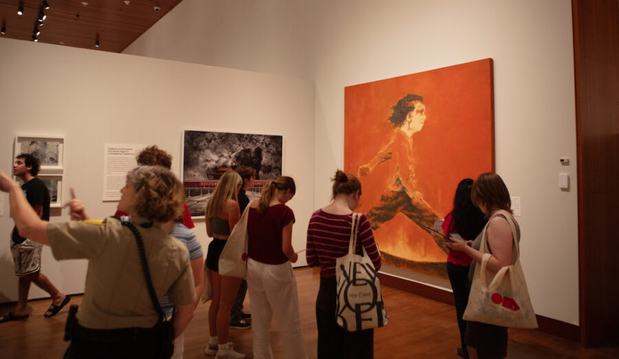 A group of people gathers in an art gallery, attentively viewing a large, vibrant painting of a figure running against a fiery orange backdrop.