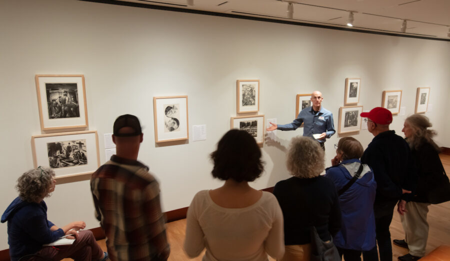 A guide gestures toward framed artworks on a gallery wall as a group of visitors attentively observes the display.