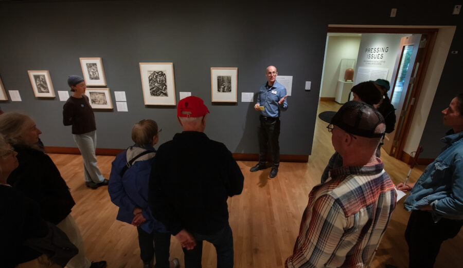 A group of people gathers in an art gallery, attentively listening to a guide who is explaining the displayed artworks.