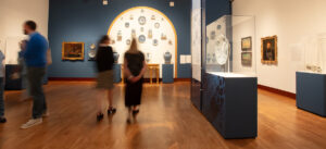 Visitors stroll through a museum gallery featuring a prominent arched display of decorative plates, framed artwork, and glass-encased artifacts on wooden floors.