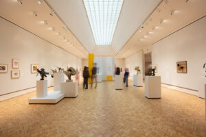 Visitors stroll through a spacious, well-lit gallery with wooden flooring, white walls displaying framed art and sculptures on pedestals, under a long skylight.