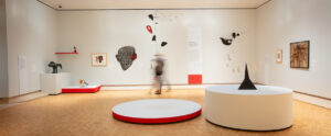 A modern art gallery with minimalist white walls, wooden flooring, and circular display platforms showcasing abstract sculptures and paintings.