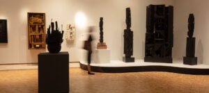 A gallery space features several dark, abstract sculptures displayed on pedestals against white walls, with a blurred figure walking through the room.