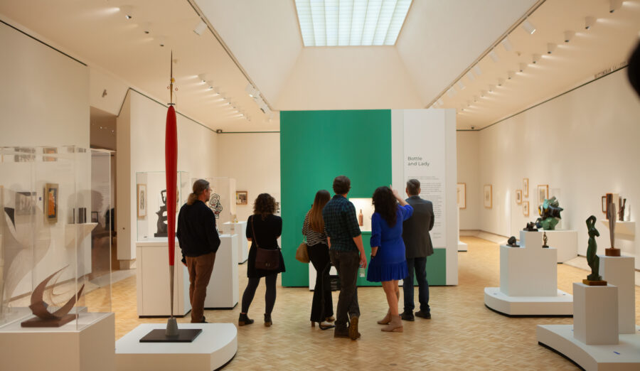Visitors explore a modern art gallery with sculptures and paintings displayed on white pedestals under bright, even lighting.