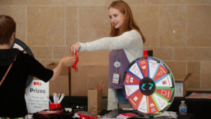 A person hands a red item to another individual at a table featuring a prize wheel and various giveaways.