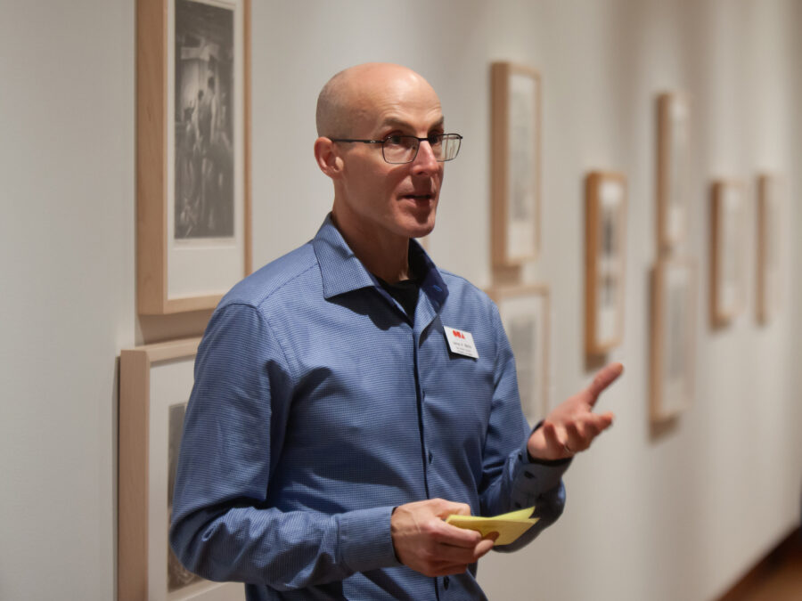 A person in a blue shirt gestures while speaking in an art gallery, with framed artworks on the walls and another individual in the background.